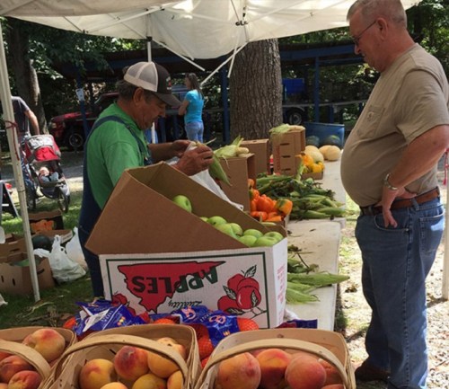 A farmer's market scene with a vendor selling fresh produce to a customer near boxes of apples and peaches.
