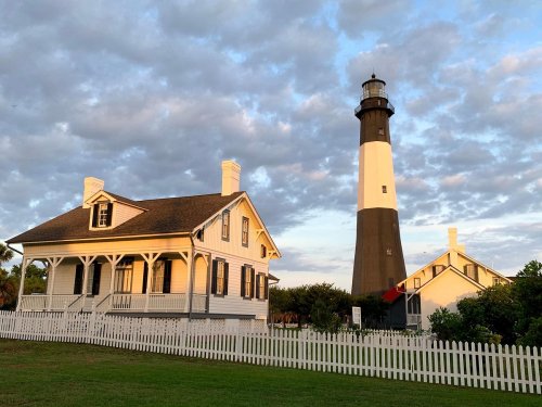 A historic lighthouse beside a white picket fence and a charming house, set against a cloudy sky at sunset.