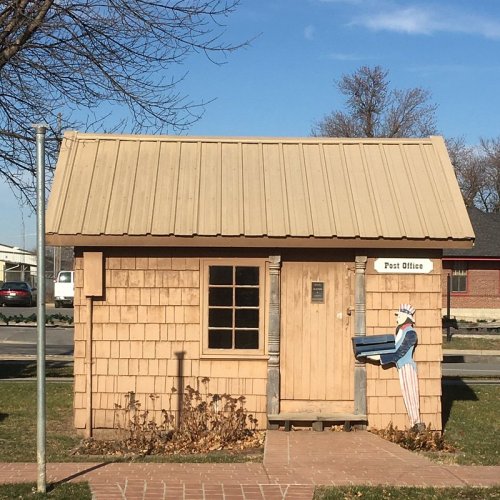 A small wooden post office building with a sloped roof and a painted figure of a mailman on the side.