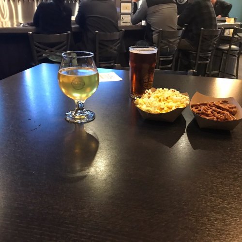 A table with a glass of beer, a bowl of popcorn, and a dish of pretzels, with people seated in the background.