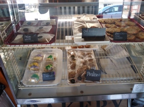 Display case filled with assorted baked goods, including cookies, pralines, and king cake slices, labeled on chalkboards.