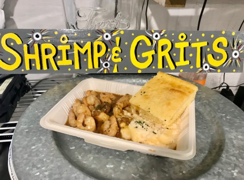 A plate of shrimp and grits with a piece of cornbread, next to a colorful sign reading "Shrimp & Grits."