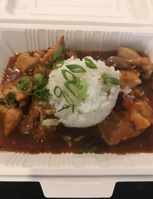 A takeout container with chicken in a savory sauce, topped with green onions and a mound of white rice.