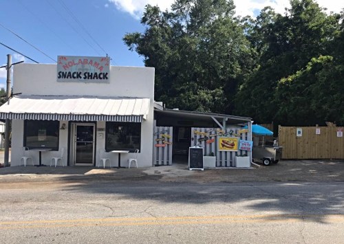 A small, white building with a striped awning, labeled "Nolabama Snack Shack," surrounded by trees and outdoor seating.