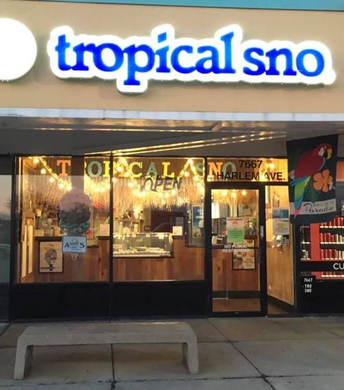 A storefront with a sign reading "tropical sno," featuring bright lights and an open sign.