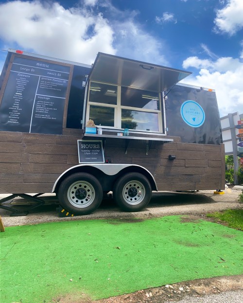 A food truck with a black exterior and menu board, parked on green grass under a blue sky with clouds.