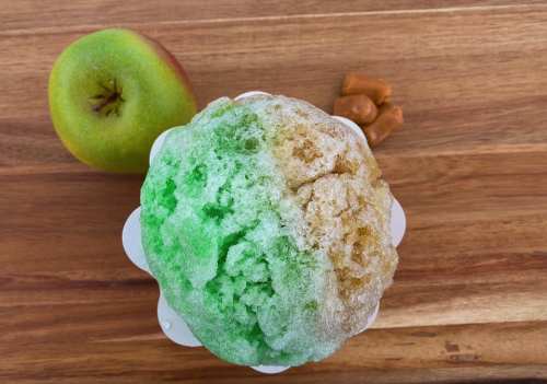 A colorful snow cone with green and brown flavors, next to a green apple and caramel candies on a wooden surface.