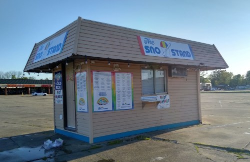 A small, colorful kiosk named "The Sno Stand" with a menu displayed outside, set in an empty parking lot.