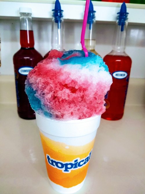 A colorful snow cone in a cup with a pink straw, surrounded by bottles of flavored syrups.