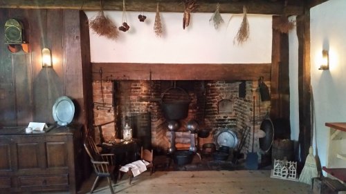 A rustic kitchen with a large brick fireplace, hanging herbs, and various cooking utensils on display.