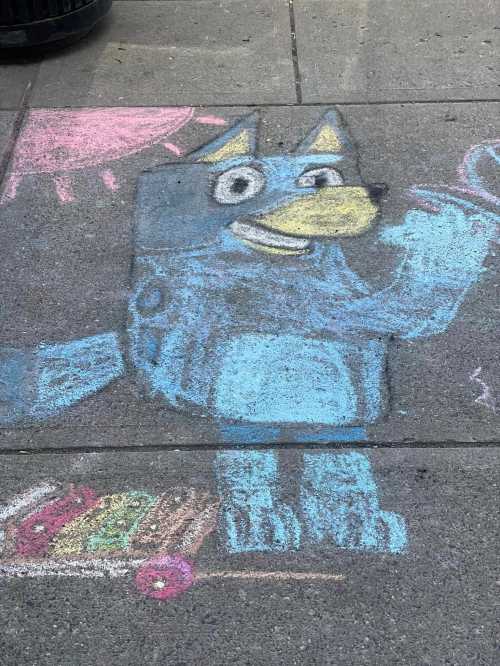 Chalk drawing of a blue cartoon wolf with a yellow beak, waving next to a colorful cart and a sun in the background.