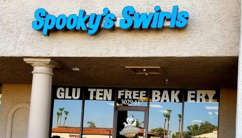 Sign for "Spooky's Swirls" gluten-free bakery, featuring blue lettering on a beige building with palm trees in the background.