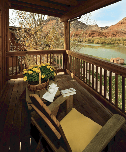 A cozy wooden porch with a chair, a basket of flowers, and a view of a river and red rock formations.