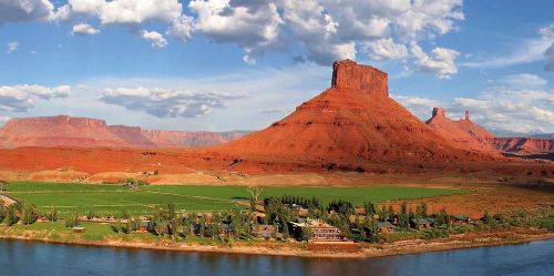 A scenic view of red rock formations and a river, with green fields and clouds in a blue sky.