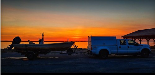 A truck towing a boat at sunset, with a calm lake and colorful sky in the background.