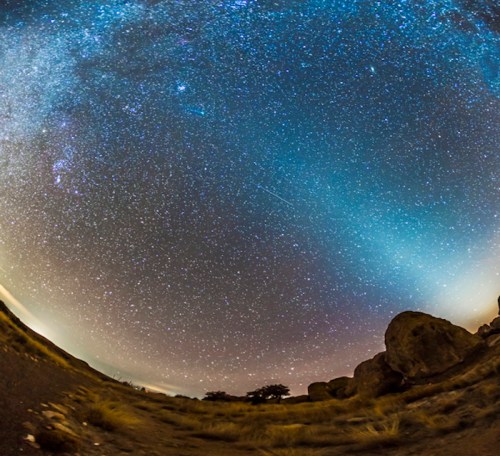 A wide-angle view of a starry night sky filled with stars and a faint glow, with rocky terrain in the foreground.