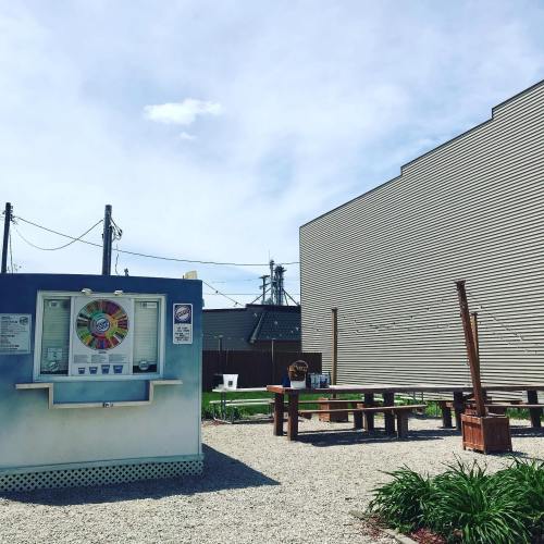 A colorful kiosk with a spinning wheel stands in a gravel area, surrounded by picnic tables and buildings under a blue sky.