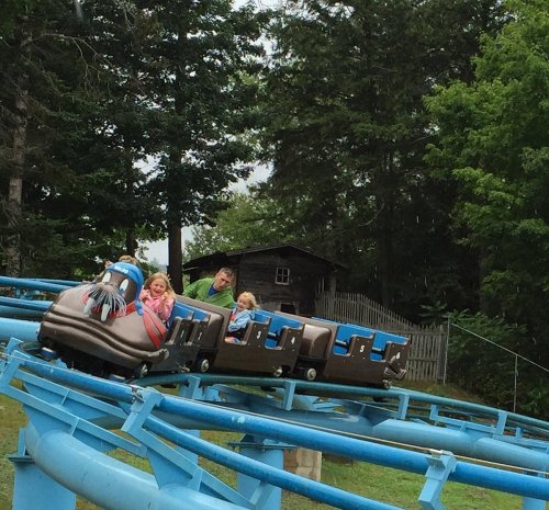 Two children and an adult enjoy a roller coaster ride, surrounded by trees and a rustic building in the background.