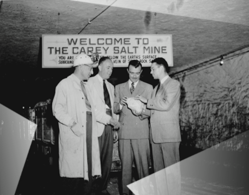 Four men in suits and coats stand under a sign that reads "Welcome to the Carey Salt Mine," discussing something together.