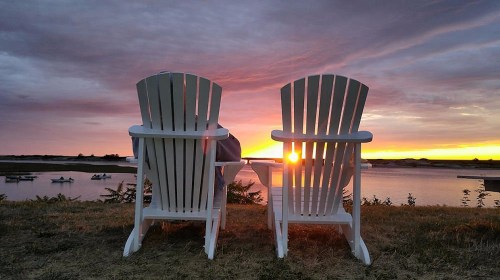 Two white adirondack chairs face a vibrant sunset over a calm river, with boats in the distance and a serene atmosphere.