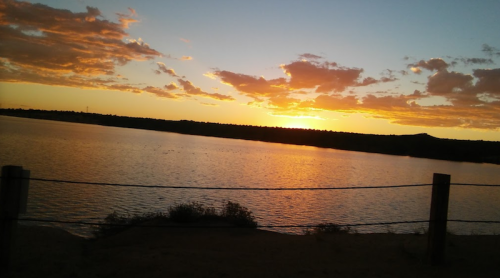 A serene sunset over a calm lake, with colorful clouds reflecting on the water and a fence in the foreground.