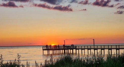 Silhouette of a fishing pier at sunset, with vibrant orange and purple skies reflecting on calm waters.