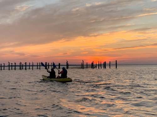 Two kayakers paddle on calm water at sunset, with silhouetted posts in the background against a colorful sky.