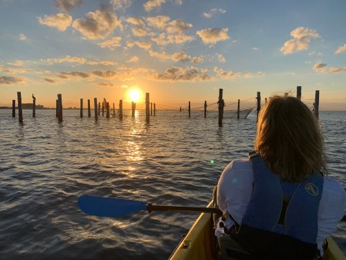 A person in a kayak watches a sunset over water, with wooden posts silhouetted against the colorful sky.