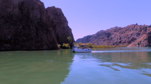 A boat glides through calm green waters surrounded by rocky cliffs and mountains under a clear blue sky.