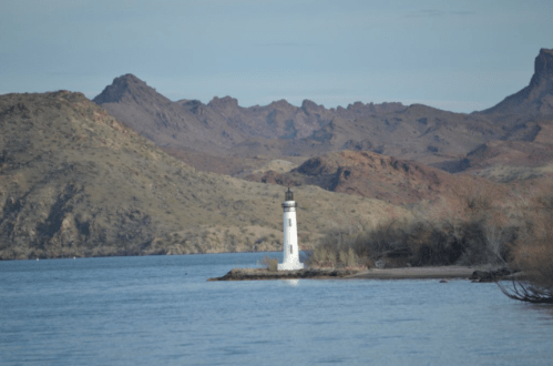 A white lighthouse stands on a small island, surrounded by calm water and rugged mountains in the background.