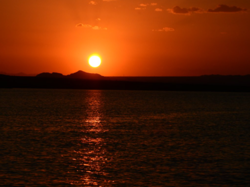 A vibrant sunset over a calm body of water, with silhouettes of distant mountains against an orange sky.