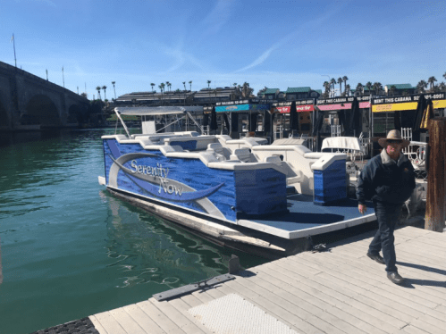 A boat named "Serenity Now" docked at a marina with a person walking nearby. Palm trees and buildings in the background.