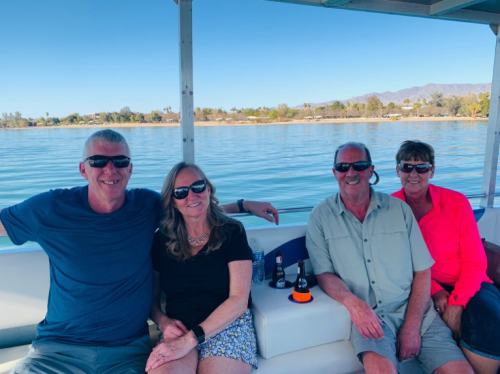 Four people sitting on a boat, smiling and enjoying a sunny day by the water with mountains in the background.