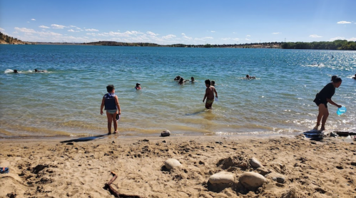 Children and adults play in the water at a sandy beach on a sunny day, with a clear blue sky and calm lake.