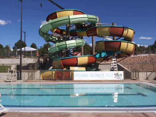 Colorful water slides tower over a clear swimming pool under a blue sky, with a sign announcing summer 2020 opening.