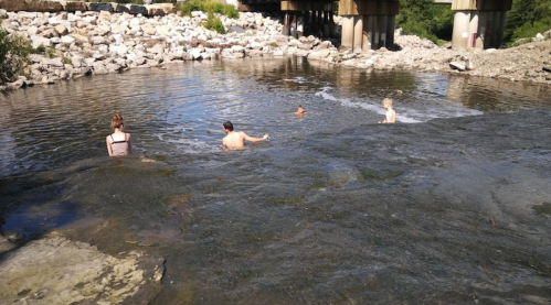 Four people wade in a shallow river under a bridge, surrounded by rocky banks and greenery.