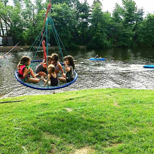 A group of children sitting on a swing by a river, enjoying a sunny day with trees in the background.