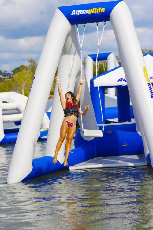 A girl in a life jacket joyfully swings from an inflatable structure over water, splashing as she jumps.