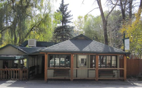 A cozy, single-story cabin with a green roof, surrounded by trees and a wooden porch.