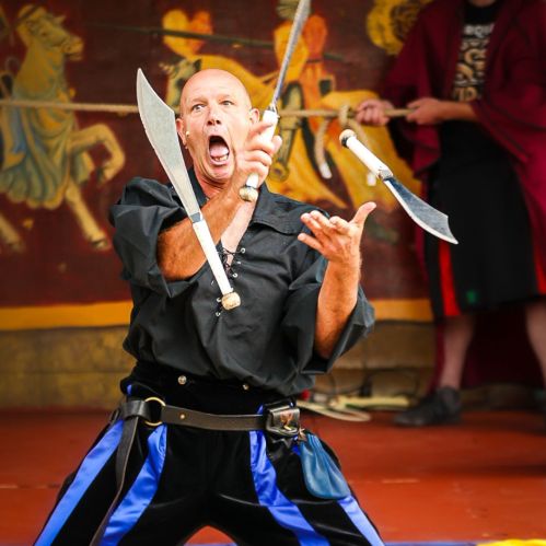 A performer in a black outfit juggles swords with an intense expression during a live show.