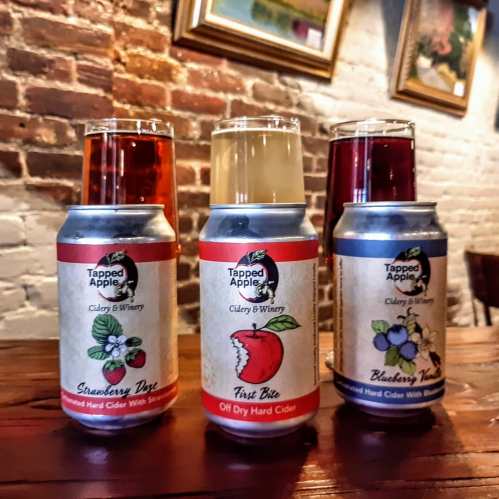 Three cans of Tapped Apple hard cider on a wooden table, with glasses of cider in the background.
