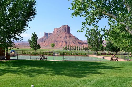 A scenic view of tennis courts surrounded by green grass and trees, with a red rock formation in the background.