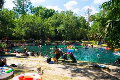 A lively swimming area with people enjoying the water, surrounded by trees and colorful floaties on a sunny day.