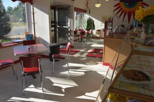 Bright café interior with red chairs, tables, and a display of baked goods. Sunlight casts shadows on the floor.