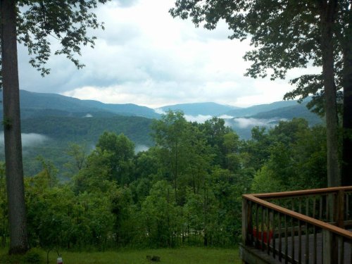 A scenic view of misty mountains and lush green trees under a cloudy sky, with a wooden railing in the foreground.