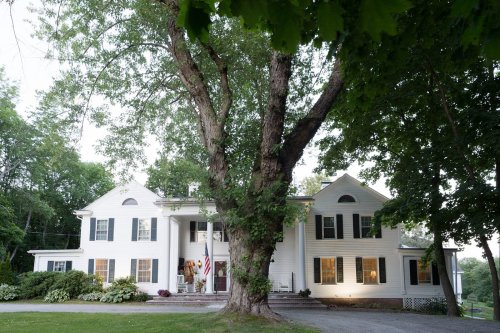 A large white house with a porch, surrounded by trees and greenery, featuring an American flag at the entrance.