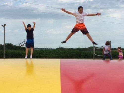 Two boys jump on a large inflatable bounce pad, with one mid-air and the other standing on the edge.