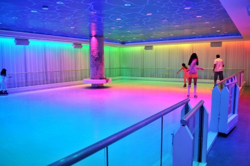 A colorful ice skating rink with vibrant lights, featuring a central pillar and skaters enjoying the space.