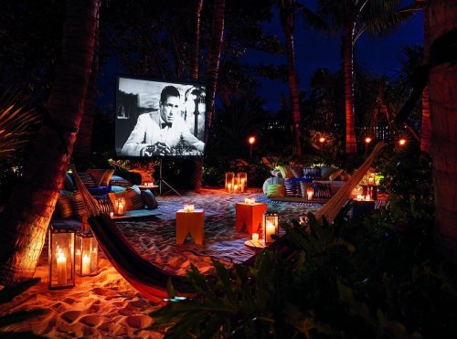 A cozy outdoor movie setup with a large screen, candles, and lounge seating surrounded by palm trees at night.