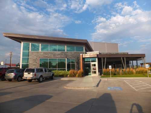 Modern building with large windows, a flat roof, and landscaped entrance, surrounded by parked cars and blue sky.